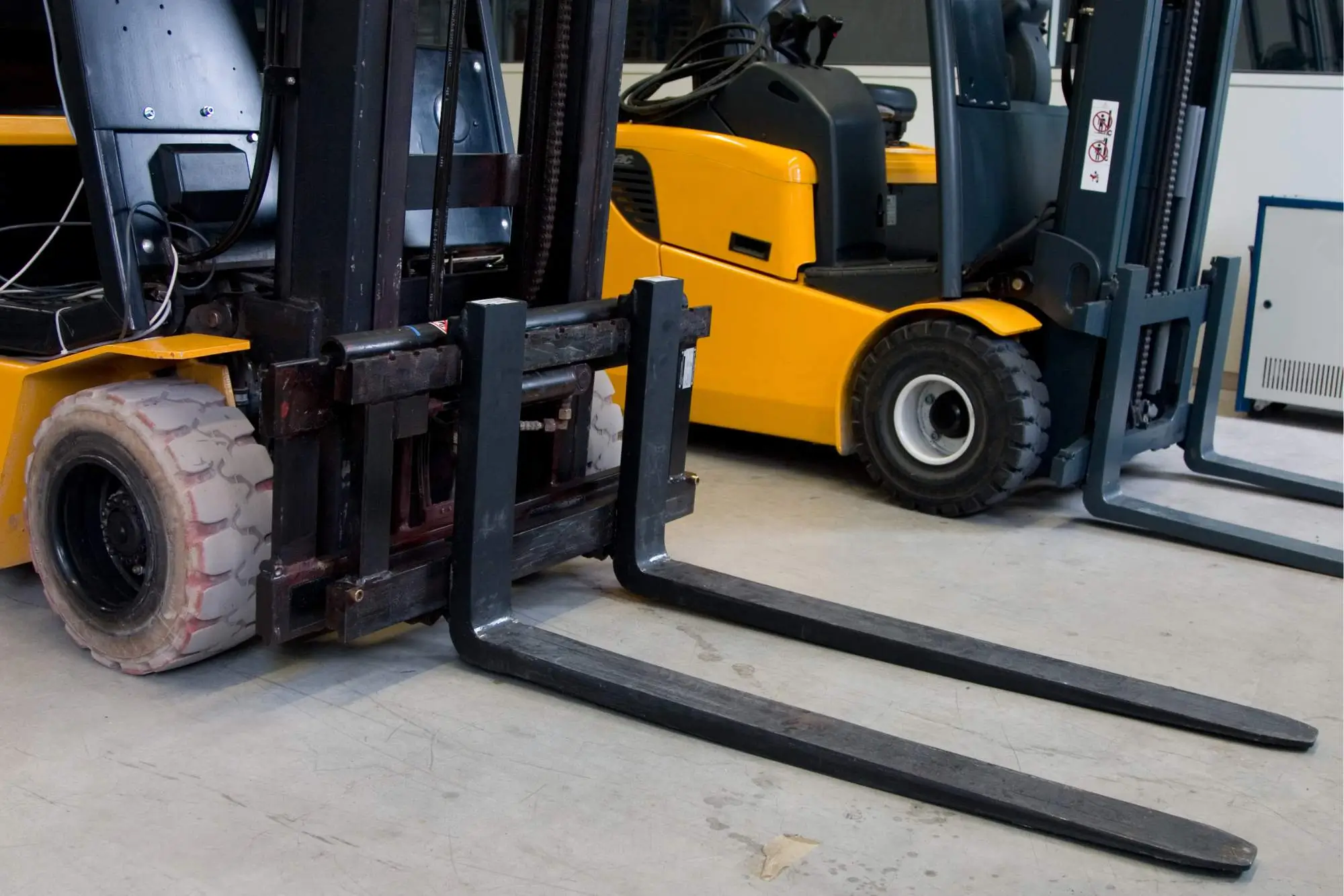 Two yellow forklifts parked indoors with their black lift forks lowered onto a concrete floor.